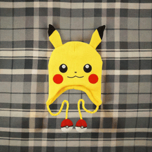 Load image into Gallery viewer, Pokemon Laplander Pikachu Beanie with Pikachu face, ears, and Poké Ball tassels on plaid background.